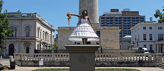 A black woman in a white dress stands on an empty pedestal of a dismantled Confederate monument