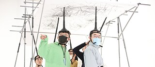 Several people are standing in the exhibition space. They carry large pens on their heads, drawing on the ceiling.