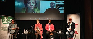 İki kadın ve iki erkek yüksek taburelerde oturuyor. Her birinin önünde bir stand üzerinde bir mikrofon bulunuyor. Arka planda, çevrimiçi bir oturumda olan iki kişinin fotoğraflarının görüldüğü bir projeksiyon var. Konuşmacıların solunda bir Festival Academy roll-up banneri ve sağında üzerinde tam görünmeyen kırmızı harfler olan beyaz bir banner görülüyor.