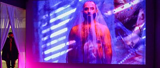 Eine Videoprojektion an einer Wand zeigt einen oben ohne Mann, der mit einem durchsichtigen Schleier bedeckt ist. Ein Teil des Gesichts des Mannes, der einen Ohrring und eine Perlenkette trägt, wird ebenfalls in den Hintergrund projiziert. Auf der linken Seite des Bildes sehen wir denselben Mann in schwarzer Kleidung zwischen zwei weißen Vorhängen stehen. 
