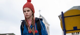 Szene aus dem Film 'Mission Ulja Funk'. Ein kleines Mädchen mit Zöpfen und einer roten Mütze mit Bommeln steht mit entschlossener Miene da.