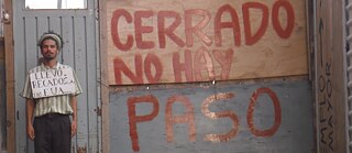 A man is standing in front of a door, on the wall next to him is written “CERRADO NO HAY PASO”.