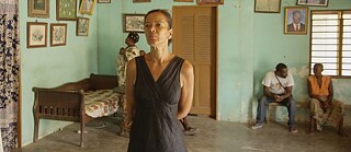 Sala de espera con personas sentadas y una mujer de pie en el primer plano. Por su aspecto, parecen ser personas africanas.