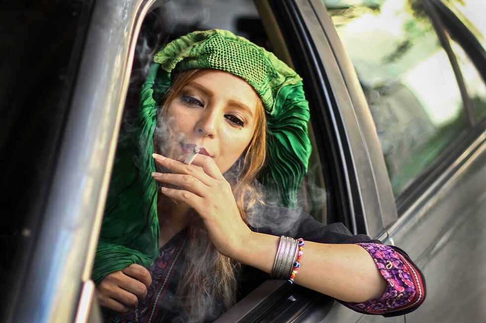 Eine Frau, die am Steuer eines Autos sitzt, ihre Burka nach hinten geschlagen hat und eine Zigarette raucht