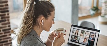 Жена држи шолја за кафе во рацете и седи пред лаптоп со слушалки во увото.