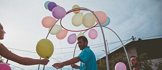  A woman hands a balloon to a man holding several colourful balloons and standing on a cart.