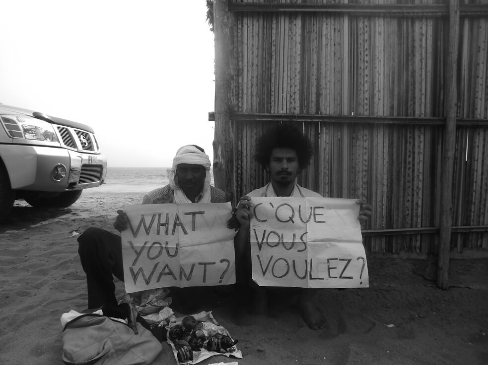 Two men sit in the desert with signs that say "what you want?". 
