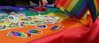 Rainbow colored stickers are laid upon a rainbow flag. In the background a hand is picking up a sticker.