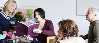 Three middle-aged women and one man are sitting in a classroom. The woman in the centre shows to the lady next to her something in a book with purple cover that she is holding. The other two persons are looking at them.
