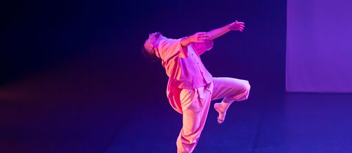 A dancer is on the stage. He is standing on his right foot and his left leg is raised. Both his arms are up in the air extended in front of him at shoulder height. His head is tilted backwards, looking towards the sky. The stage looks dark and a pink light is washing over him.