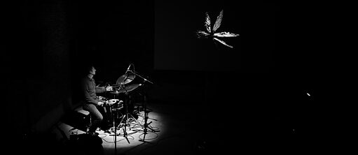 The black and white photo is mainly dark. In the left side of the picture, a drummer is illuminated, sitting in front of his drum kit. His face is turned away from the camera. In the upper right part of the picture you can see the contours of a butterfly.