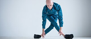 Ein Tänzer steht mit gekreuzten Beinen, nach vorne gelehnt und mit hängenden Armen, die fast den Boden berühren. Er trägt ein blau-hellblau gestreiftes Kostüm und schwarze Turnschuhe. Der Boden und der Hintergrund sind weiß.