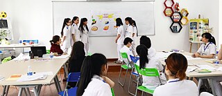 Eine Gruppe Schülerinnen hält in einem Klassenraum eine Präsentation an der Tafel. Alle tragen Pflegekräfte-Kleidung.