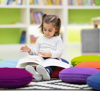 Kleines Mädchen blättert in einem Buch, sitzt auf bunten Kissen