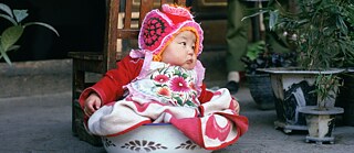 En baby sidder i en potte med en farverig kjole.  © Filmstill fra China - die Künste - der Alltag. ©Ulrike Ottinger Filmproduktion/SFB/WDR China - die Künste - der Alltag. 