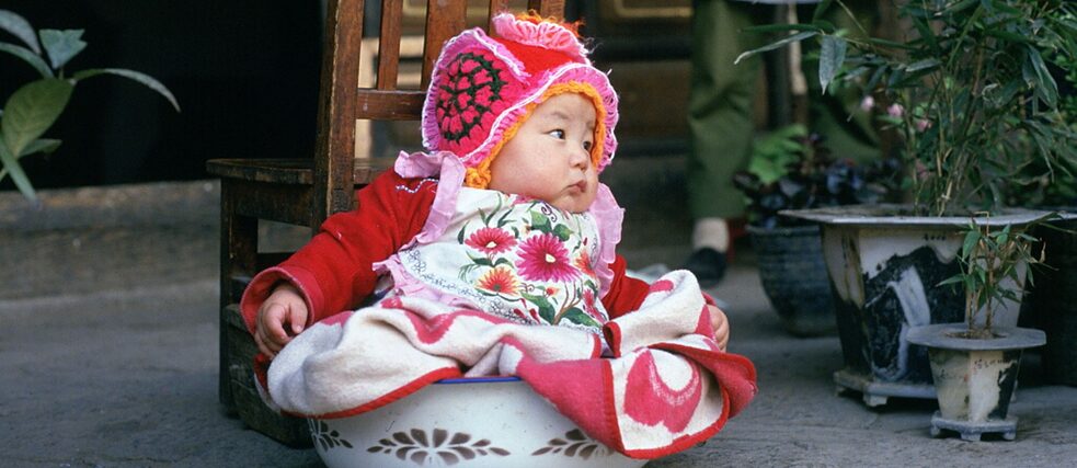 Ein Baby sitzt mit buntem Kleid in einem Topf.