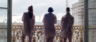 Three people on a balcony can be seen from behind, the photographic perspective is from inside a building. The people are wearing pink uniforms and white shoes while standing in front of a golden, ornamental old-fashioned balcony railing. Standing there, they are facing the city, which is the background of the picture. There, several construction sites can be made out by means of cranes and scaffolding, the sky is densely clouded.