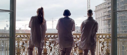 Bir balkonda duran  üç kişinin arkadan görüntüsü. Fotoğraftaki bakış açısı bir binanın içinden dışarıya doğru. İnsanlar pembe üniformalar ve beyaz ayakkabılar giymiş, altın renkli, antik tarzda süslü bir balkon korkuluğunun önünde duruyorlar. Balkondan, resmin arka planını oluşturan şehre bakıyorlar. Arkada görünen vinçler ve iskeleler birkaç inşaat alanının fark edilmesini sağlıyor. Gökyüzü çok bulutlu.