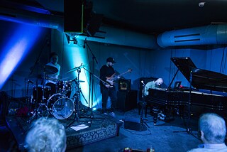 Im dunklen, blau beleuchteten Club sitzt links der Schlagzeuger Alex Poliakov, in der Mitte steht Bassist Eugene Myrmyr und rechts sitzt Andrew Pokaz am Flügel.