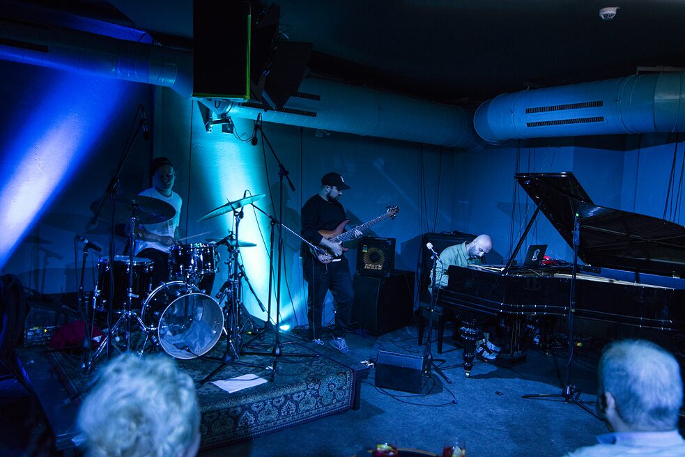 Im dunklen, blau beleuchteten Club sitzt links der Schlagzeuger Alex Poliakov, in der Mitte steht Bassist Eugene Myrmyr und rechts sitzt Andrew Pokaz am Flügel.