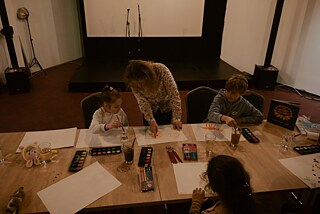 An einem großen Tisch malen drei Kinder mit Wasserfarben. Lana Ra steht in der Mitte und zeigt dem Kind links neben ihr Etwas.