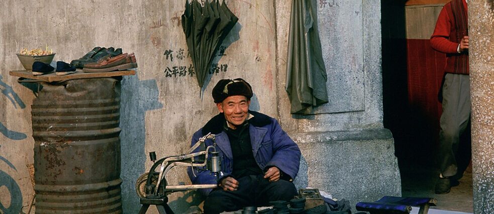 Ein lachender Mann sitzt vor einer Häuserwand, er repariert Schuhe. 