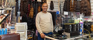 Ein Mann steht hinter einer Ladentheke und lächelt in die Kamera. Hinter ihm hängen mehrere Haarteile zum Verkauf aus.