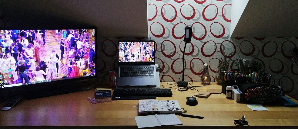 Stůl s obrazovkou, notebookem a psacími potřebami.