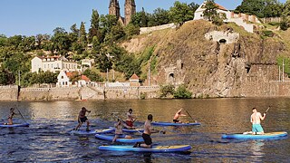 Auf dem Foto ist der Prager Burgwall Vyšehrad zu sehen. Im Vordergrund fließt die Moldau, auf welcher einige paddelnde Personen auf Surfbrettern zu sehen sind. Im Hintergrund, hinter dem Wall, sind Felsen, Bäume und eine Kirche in strahlendem Sonnenschein zu sehen.