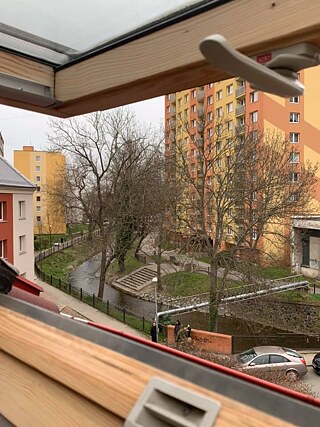 Ich lebe mit meiner Familie in einer kleinen Stadt in der Nähe von Prag in einem Familienhaus. Mein Zimmer ist auf dem Dachboden und das ist meine Aussicht. Vom Fenster aus sehe ich einen Bach, Wohnblöcke und einen der örtlichen Kindergärten. Meine jetzige Schule ist in der Nähe. Es ist buchstäblich um die Ecke. Ich kann sie vom Balkon aus sehen. Es gibt viele Schulen in der Stadt. Es gibt 4 Supermärkte und unzählige Lebensmittel in der Stadt. Es gibt auch Cafés, Restaurants, Casinos, Pubs… Es gibt auch einen Bahnhof, ein Kino, eine Kirche usw.
