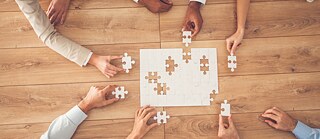Několik rukou společně skládá velké puzzle na dřevěném stole, symbol spolupráce a týmové práce při výuce němčiny.