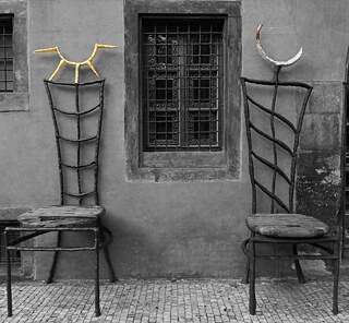 Auf dem Foto sind zwei dunkelgraue Stühle zu sehen. Am linken Stuhl ist oben eine halbe goldene Sonne befestigt, am rechten Stuhl ein silberner Halbmond. Sie stehen auf Kopfsteinpflaster und vor einer grauen Wand. Zwischen den Stühlen ist ein schwarz vergittertes Fenster in der Wand. 