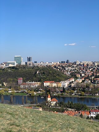 Auf dem Foto ist ein Ausblick auf Prag zu sehen. Die obere Hälfte des Fotos zeigt strahlend blauen Himmel, darunter sind zahlreiche Häuser und Hochhäuser, sowie Parks zu sehen. Im unteren Bereich des Bildes ist die Moldau zu sehen.