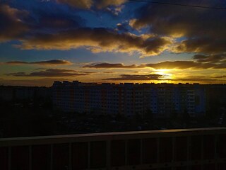 Das Bild zeigt einen Sonnenuntergang. Im oberen Teil des Bildes ist ein bewölkter Himmel zu sehen, der von einem Blau oben in ein Gelb am Horizont verläuft. Im unteren Bereich des Bildes liegt der Stadtteil Stodůlky im Schatten.