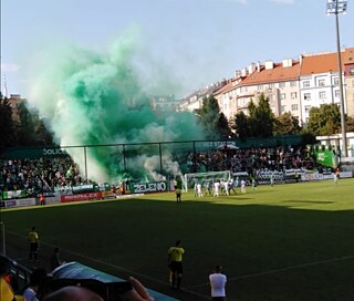 Das Foto zeigt das Stadion Ďolíček. Im unteren Bereich des Fotos ist grüner Fußballrasen mit Spieler:innen in weißen und grünen Trikots zu sehen. Im oberen Bereich des Fotos ist die Tribüne zu sehen, von der aus grüner Rauch aufsteigt. Im Hintergrund sind außerdem einige Häuser und blauer Himmel zu sehen.
