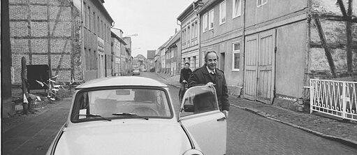 Mann steht neben seinem Auto, ein Käfer, mit offener Tür, auf einer Dorfstraße. Die Aufnahme ist in schwarz-weiß.