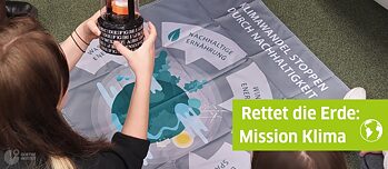 Ein Mädchen sitzt vor einem Plakat, auf dem verschiedene Möglichkeiten der nachhaltigen Energiegewinnung abgebildet sind. In seinen Händen hält es einen Tresor mit Ziffernschloss.