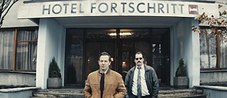Dos hombres de pie frente al acceso a un hotel cuyo nombre aparece escrito sobre la marquesina y dice Hotel Fortschritt (Hotel del progreso)