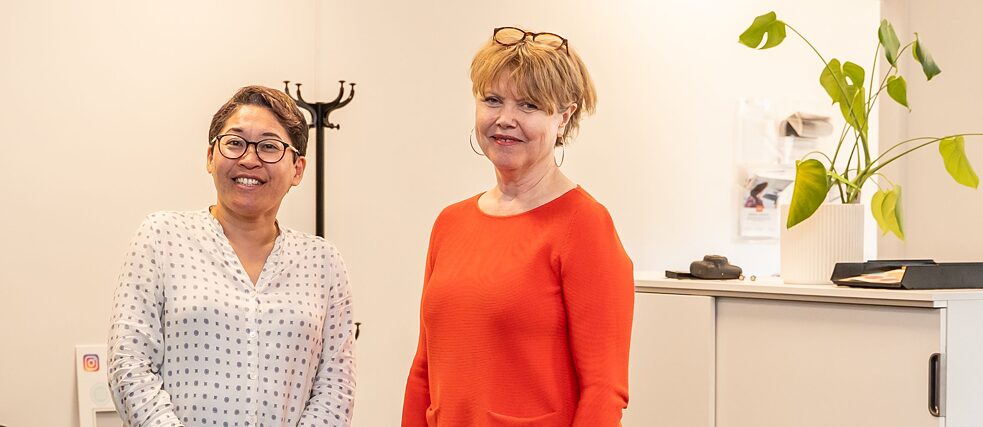 Zwei Frauen stehen in in einem Büro-Raum. Sie tragen beide Brillen und blicken direkt in die Kamera.