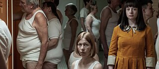 Eine Gruppe von Erwachsenen steht in weißer Unterwäsche in einer Umkleidekabine. In der Bildmitte eine sitzt eine Frau und schaut in die Kamera. Am rechten Bildrand steht die Hauptdarstellerin in einem gelben Kleid und schaut ebenfalls in die Kamera. © © The Match Factory The Ordinaries