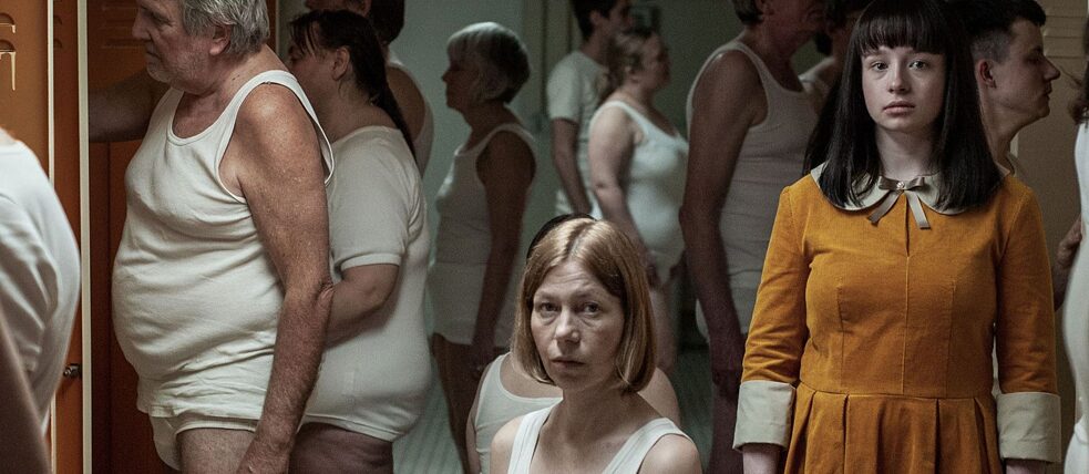 Eine Gruppe von Erwachsenen steht in weißer Unterwäsche in einer Umkleidekabine. In der Bildmitte eine sitzt eine Frau und schaut in die Kamera. Am rechten Bildrand steht die Hauptdarstellerin in einem gelben Kleid und schaut ebenfalls in die Kamera.