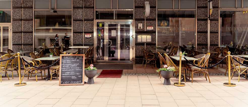 Außenansicht eines Cafés. Mehrere Tische stehen im Freien vor einer Glasfront.