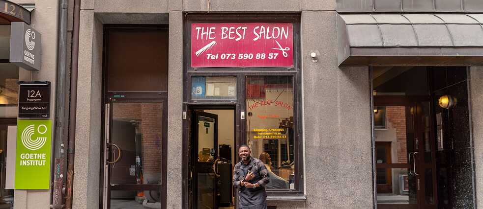 Außenansicht eines Friseursalons. Pappa Matha, der Besitzer, steht davor und lächelt in die Kamera.