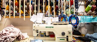 Eine Nähmaschine steht auf einem Tisch. Daneben liegen Klamotten. Darüber befindet sich ein Regal mit verschiedenfarbigem Garn.