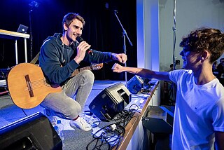 Ein Musiker mit Gitarre und Mikrofon auf der Bühne hockt und gibt Kind vor der Bühne einen Fistbump.