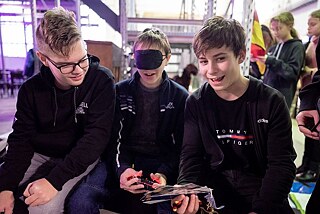 Drei Jungen, der mittlere hat verbundene Augen und eine Fernsteuerung in der Hand, sie lachen.