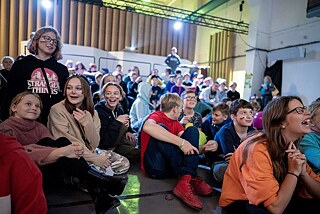 Eine große Gruppe Kinder sitzt auf dem Boden und betrachten etwas hinter der Kamera, sie lachen.