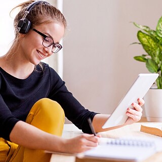 Junge Frau mit Kopfhörern sitzt am Tisch, hält in einer Hand ihren Tablett-PC und schreibt in ihren Notizblock