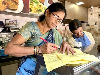 Eine Frau mit Brille steht am Tresen eines Imbiss und schreibt auf Dokumenten mit einem Stift