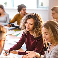 Eine junge Frau sitzt umgeben von Leuten in einem Klassenzimmer. Sie lächelt ihr Gegenüber an.